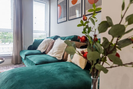 The image shows a modern styled living area with an L-shaped emerald green sofa with abstract artwork hanging behind. high-quality short term accommodation