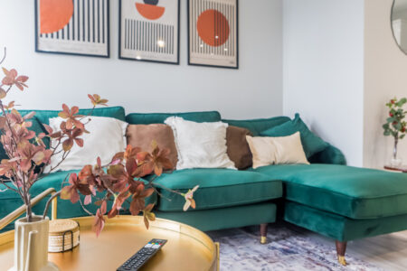 The image shows a modern styled living area with an L-shaped emerald green sofa with abstract artwork hanging behind. high-quality short term accommodation